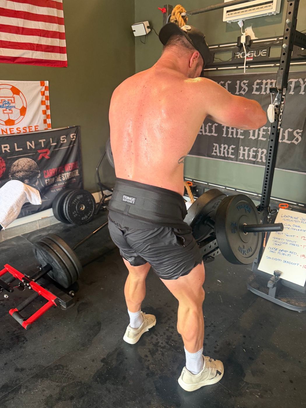 A man in a lifting belt at the landmine, next to the day's whiteboard